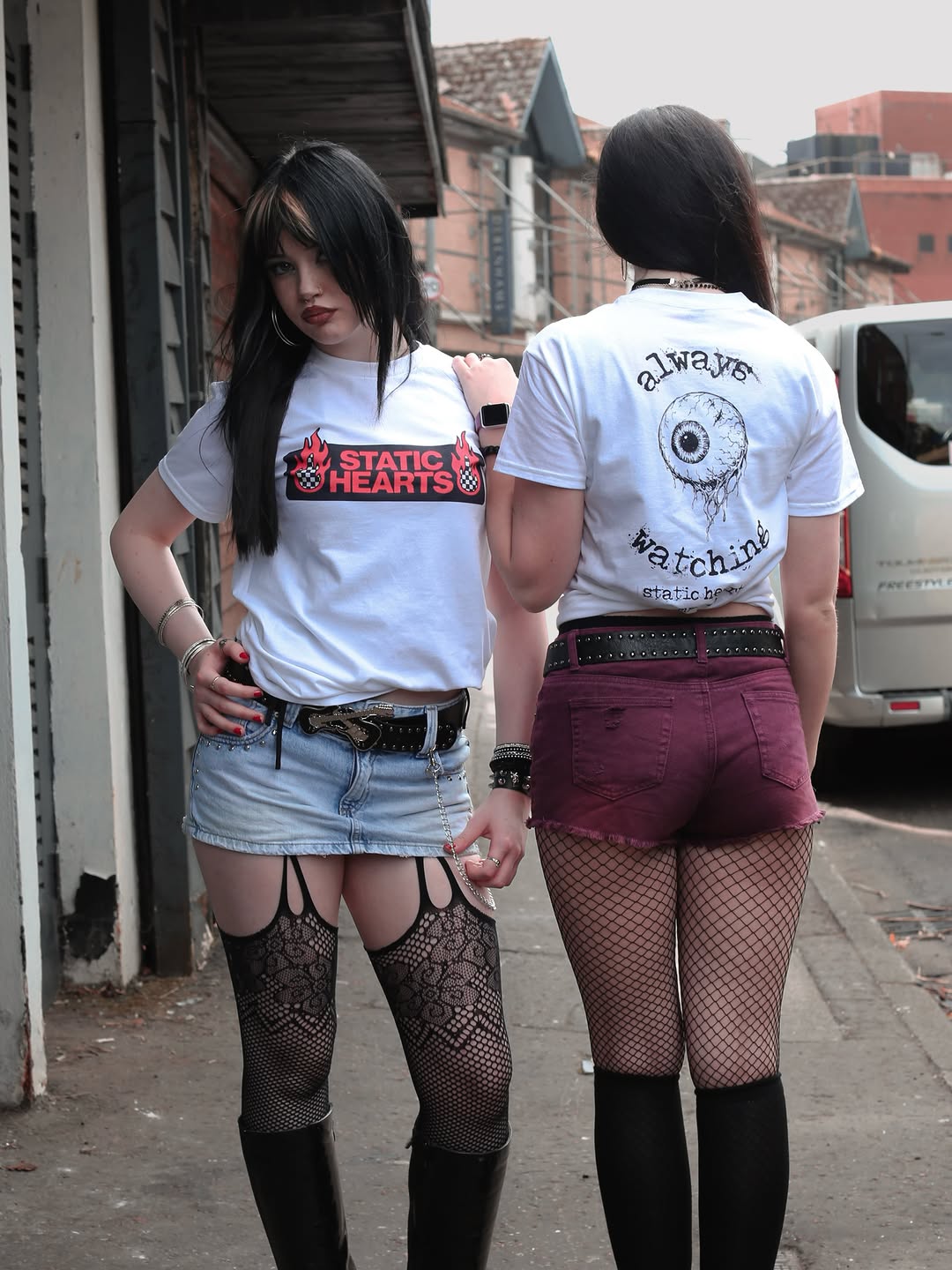 Two women in white graphic T shirts and fishnet stockings stand on a city sidewalk near a parked van