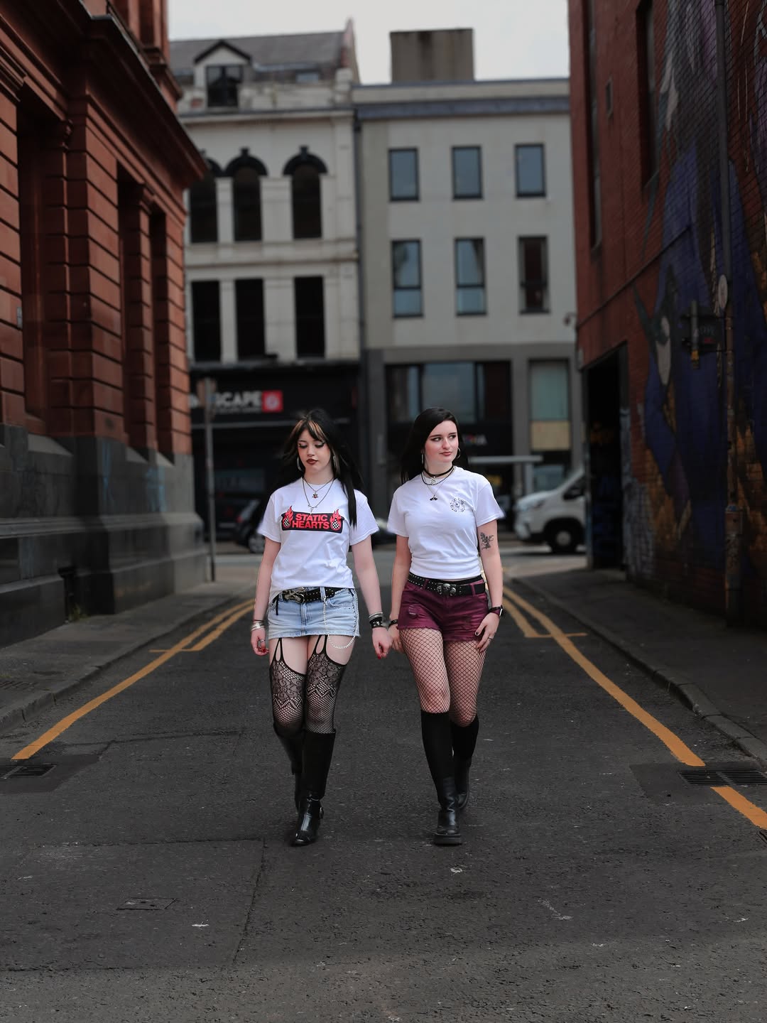 Two women walking down a narrow alley in white graphic shirts, fishnet tights, and black boots
