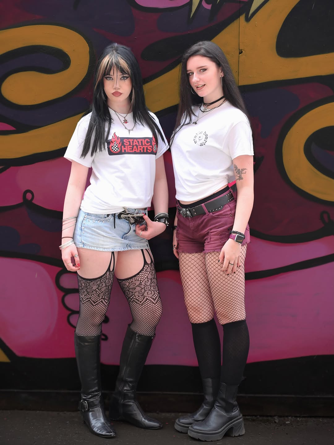 Two women in white graphic T shirts and fishnet stockings pose against a colorful graffiti wall, outdoors.